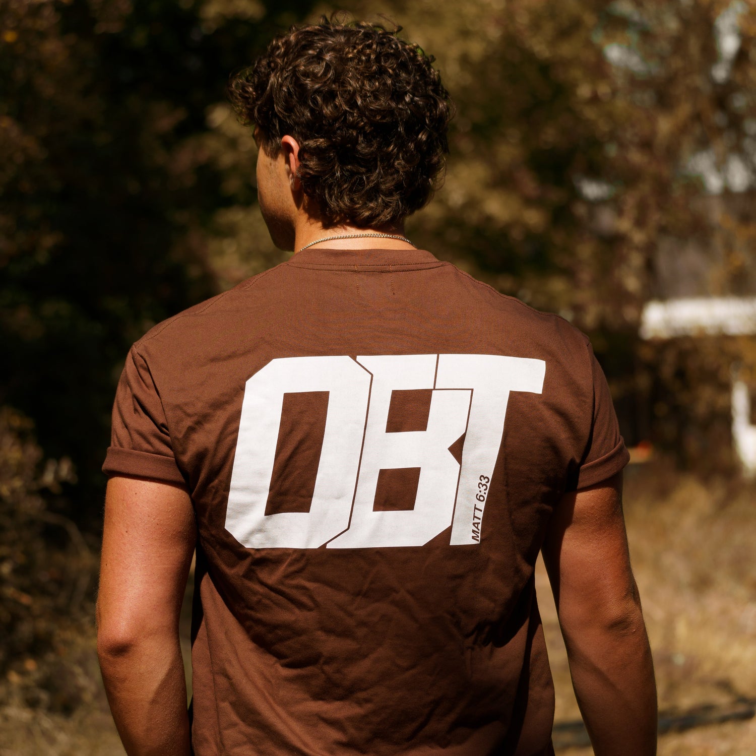 Person wearing a brown t-shirt with 'OBT' printed on the back, standing outdoors.